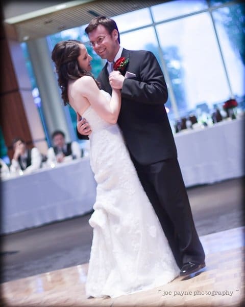 justin and meredith get hitched A bride and groom share their first dance at a wedding reception. The bride wears a white gown, and the groom is in a black suit. They smile at each other, with a blurred background of guests seated at tables.