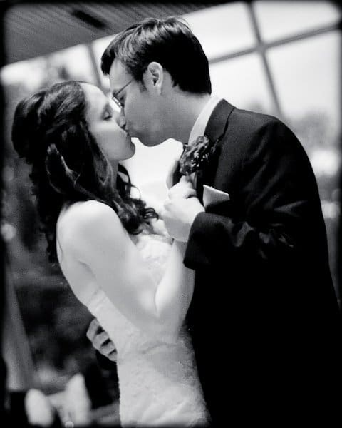 justin and meredith get hitched A black-and-white photo of a couple in formal attire sharing a kiss. The woman wears a strapless dress, and the man is in a suit with a boutonniere. They embrace intimately, set against a softly blurred background.