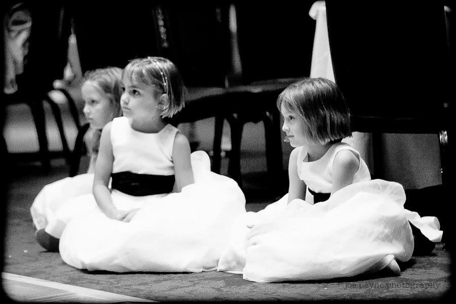 justin and meredith get hitched Three young girls in white dresses with dark sashes sit on the floor during an event. They appear attentive and are sitting in a line, with two chairs visible in the background. The image is in black and white.