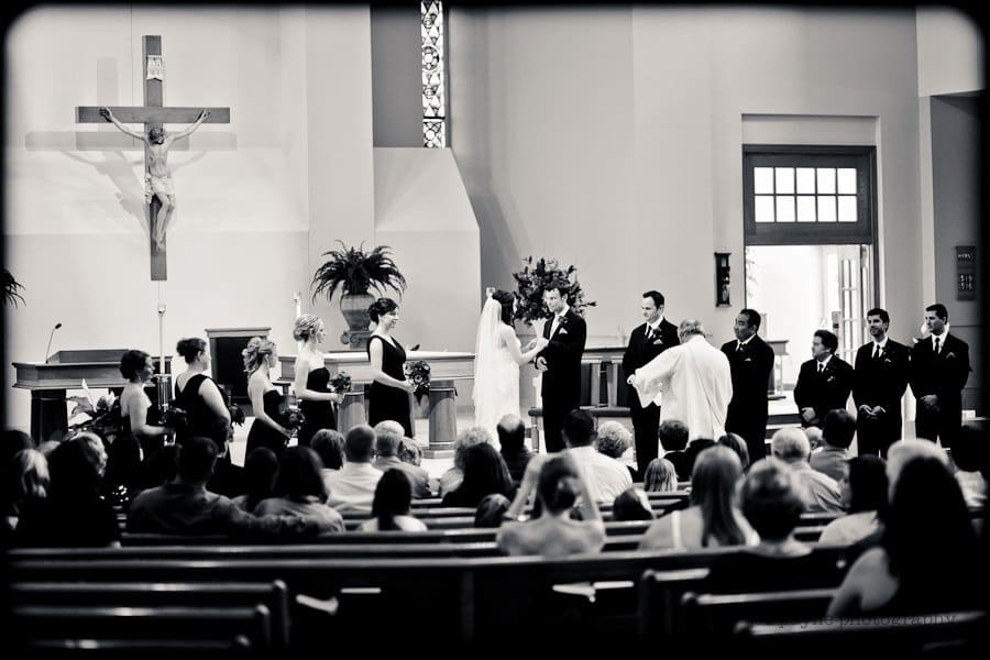 justin and meredith get hitched A black and white photo of a wedding ceremony in a church. The bride and groom stand at the altar, with bridesmaids and groomsmen on either side. A priest officiates the ceremony. Guests are seated in pews, observing the scene.