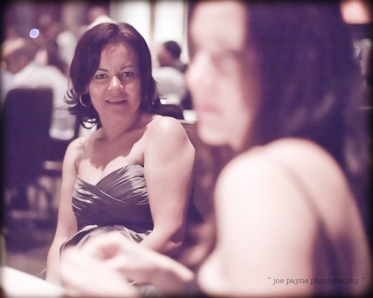 justin and meredith get hitched A woman in a strapless dress smiles softly while seated at a table in a dimly lit setting. Another person in the foreground is out of focus. The atmosphere appears intimate and relaxed.