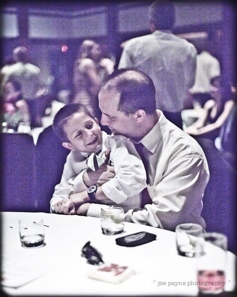 justin and meredith get hitched A man in a white shirt and tie lovingly embraces a smiling young boy at a table during a gathering. The surroundings are dimly lit with blurred people and decor in the background.