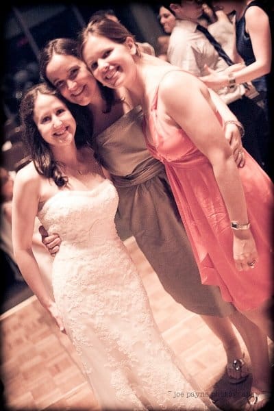 justin and meredith get hitched Three women are smiling and posing together on a dance floor. The woman on the left is in a wedding dress, while the other two are in a gray dress and a coral dress. People are dancing in the background.