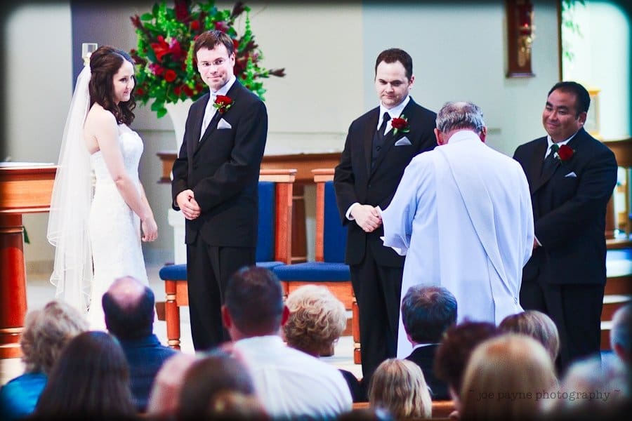 justin and meredith get hitched A bride and groom stand at the altar during their wedding ceremony, accompanied by two groomsmen. The officiant is facing them. The audience is seated in pews, and floral decorations are visible in the background.