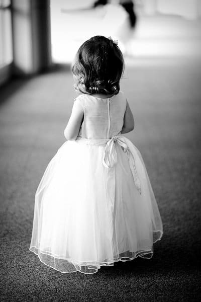 justin and meredith get hitched A black and white photo of a young girl in a white, puffy dress, standing and facing away. Her curly hair is visible from the back. The scene is softly lit, with a blurred background and flooring that looks like carpet.