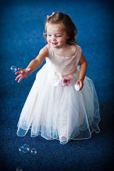 justin and meredith get hitched A young girl in a pink and white dress with floral details reaches out playfully towards bubbles in a room with a blue carpet. She has brown hair and a joyful expression.