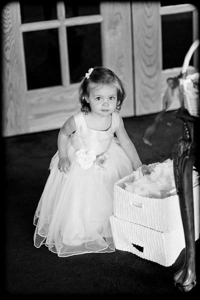 justin and meredith get hitched A young child in a formal dress with flower details stands near woven baskets. The setting appears to be indoors with wooden doors in the background. The photo is in black and white.