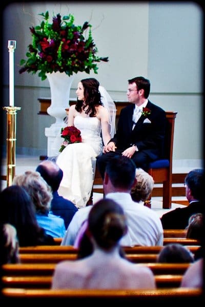 justin and meredith get hitched A bride and groom sit next to each other during a wedding ceremony in a church. The bride is holding a bouquet of red roses. A large floral arrangement is visible in the background, and guests are seated in the foreground.