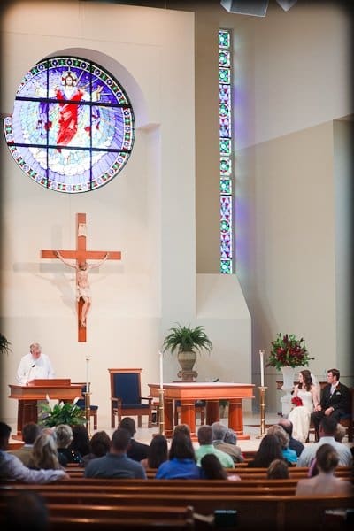 justin and meredith get hitched A church interior with a couple seated near the altar during a ceremony. A priest stands at a lectern beneath a large crucifix and a colorful stained-glass window. Congregants are seated in pews, and potted plants adorn the space.