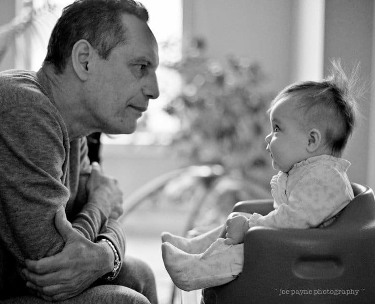 A grayscale image of an adult and a baby. The adult, sitting with arms crossed, leans in to gaze at the baby. The baby, seated in a chair, looks back with curiosity. A soft-focus background and natural lighting create a warm, intimate atmosphere.