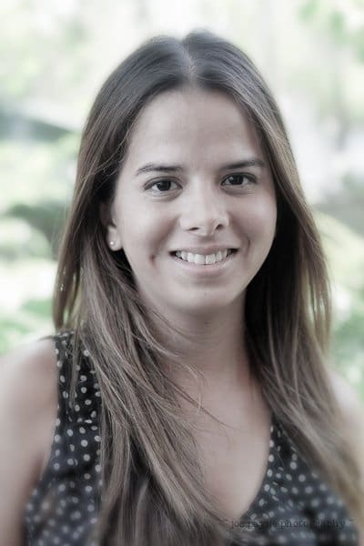 A woman with long brown hair smiles at the camera. She is wearing a black top with white polka dots. The background is blurred greenery, suggesting an outdoor setting.
