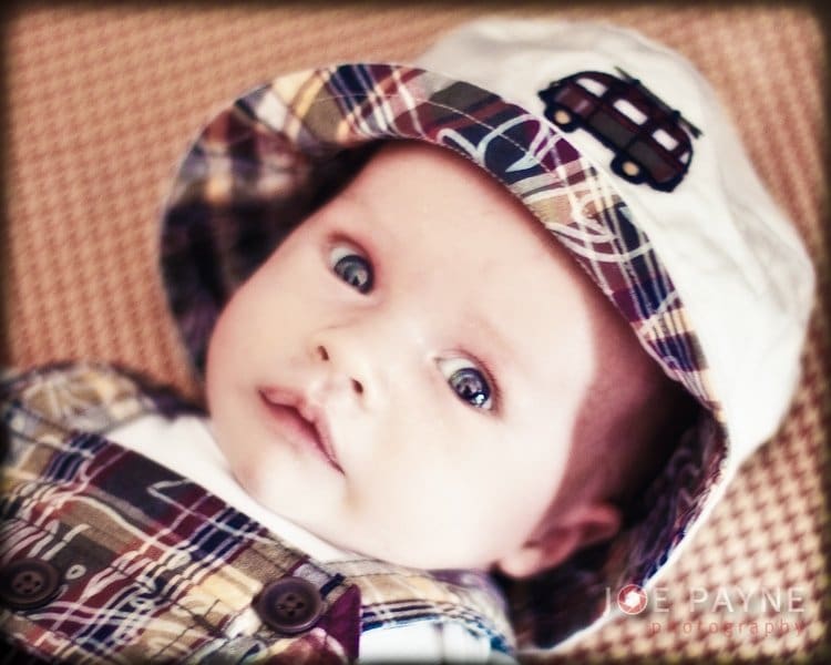 A baby wearing a plaid hat and matching overalls looks into the camera. The background is blurred, and the text JOE PAYNE Photography is visible in the corner.