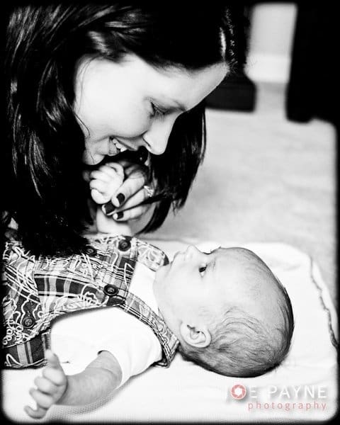 A woman with long dark hair leans in smiling at a baby lying on a blanket. The baby, in patterned overalls, looks back up at her. The atmosphere is warm and affectionate. The image is in black and white.