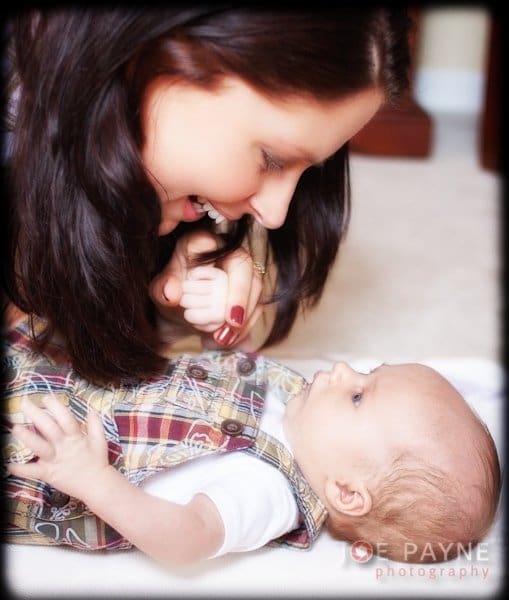 A woman with dark hair lovingly gazes at a baby lying on their back. The baby is wearing a plaid outfit. They are indoors on a soft surface, and the woman holds the babys hand gently.