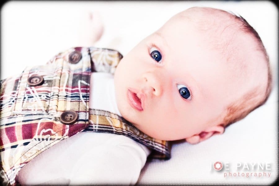 A baby with blue eyes lies on a white surface, wearing a plaid outfit with brown and red tones. The image has a soft focus effect.