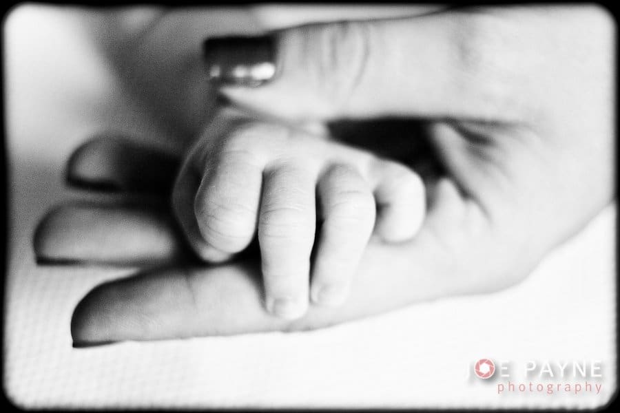Black and white photo of a babys hand gently gripping an adults finger. The scene conveys a sense of tenderness and connection.