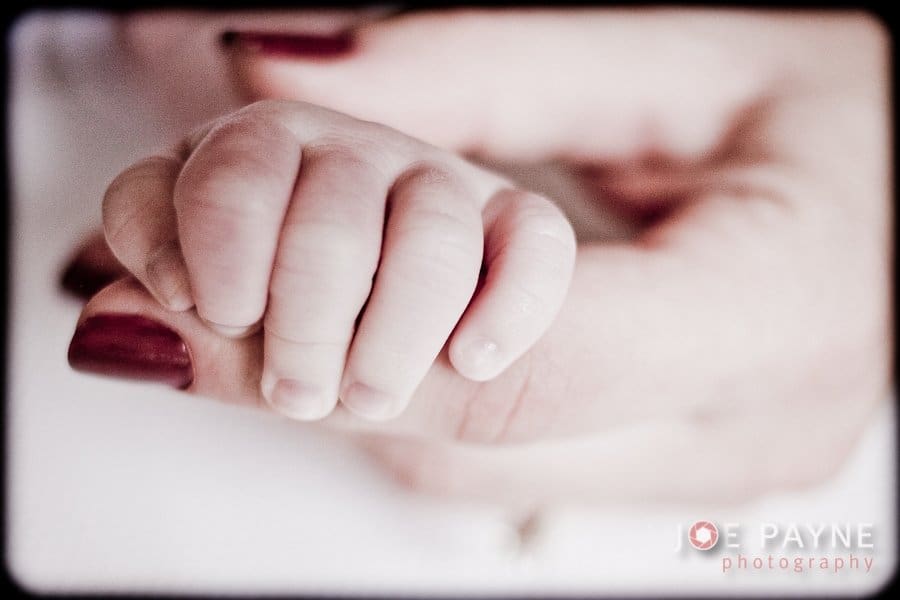 A close-up image of a babys hand gently grasping an adults finger. The adult has dark red nail polish, and the background is softly blurred, highlighting the tender moment.