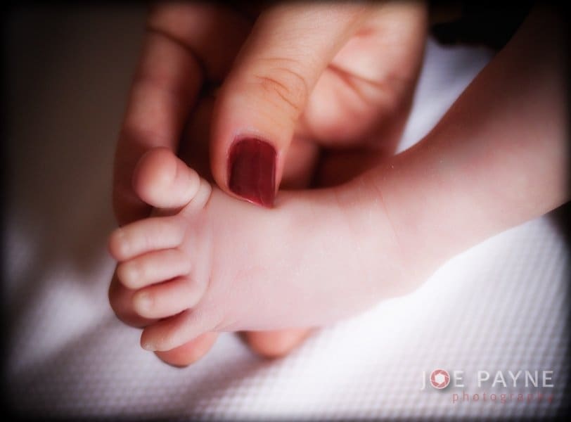 A close-up of an adult hand gently holding an infants foot. The adult has dark red nail polish, and the babys foot rests on a soft white surface. The image conveys a sense of tenderness and care.