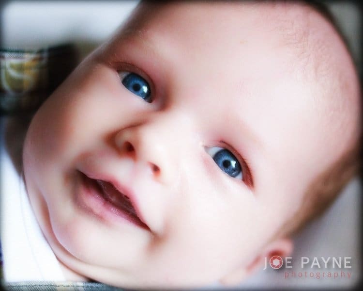 Close-up of an infant with bright blue eyes, smiling gently. The background is softly blurred, focusing on their face. The image has the photographers watermark in the corner.