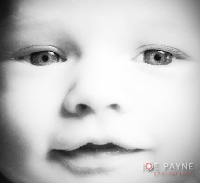 Close-up black and white photo of a babys face, showing the eyes, nose, and mouth in soft focus. The image has a gentle, serene expression. Photographers name is visible at the bottom right.