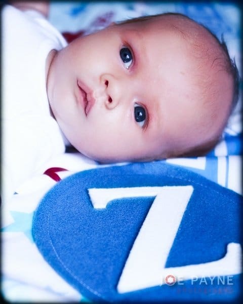 A baby lies on a soft surface, looking upward with wide eyes. Beside the baby is a blue circle with the number 7 in white. The scene has a gentle, serene atmosphere.