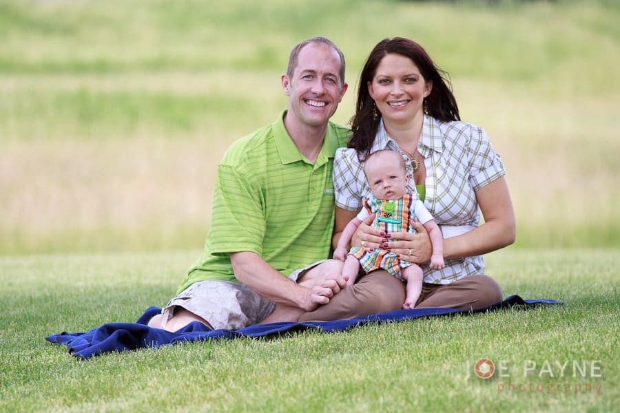 A couple sits on a blanket in a grassy field, holding a baby between them. The man is wearing a green shirt, and the woman is in a plaid top. The baby is dressed in colorful plaid overalls. They are smiling and enjoying the outdoors.