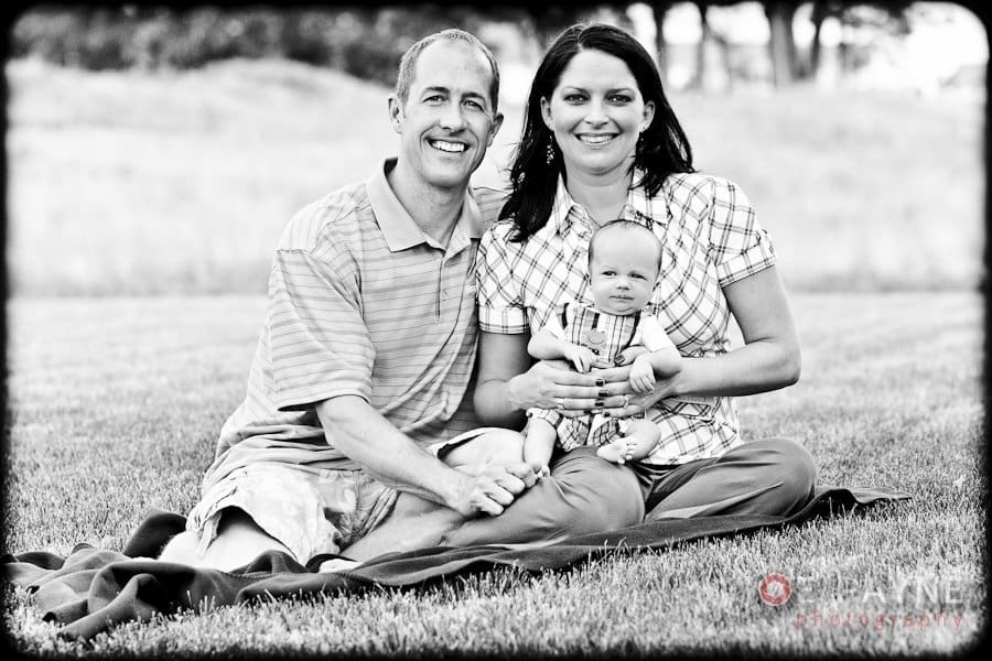 A black and white photo of a couple sitting on a blanket in a grassy field, smiling at the camera. The man wears a striped shirt and the woman wears a plaid shirt. They hold a baby dressed in overalls between them. Trees are visible in the background.