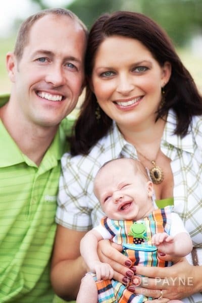 A smiling couple holds their cheerful baby. The man wears a green polo shirt, and the woman wears a plaid shirt. The baby is dressed in a colorful striped outfit with a small frog design. They are outdoors, with a blurred background.