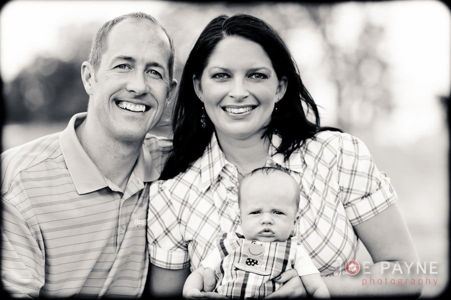 A black and white portrait of a smiling couple holding a baby. The man is on the left, wearing a polo shirt, and the woman is on the right in a checkered blouse. The baby is in the center, dressed in a plaid outfit.