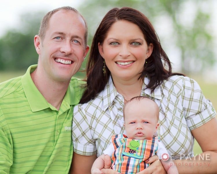 A smiling couple holds a baby outdoors. The man wears a green shirt, and the woman wears a plaid shirt. The baby is dressed in a colorful bib. The background is blurred with greenery.