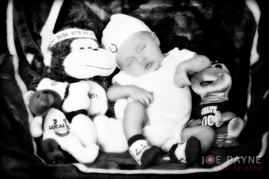 A baby wearing a beanie sleeps peacefully in black and white. The baby is surrounded by plush toys, including a monkey wearing a Dude With Attitude hat and another toy in sports attire.
