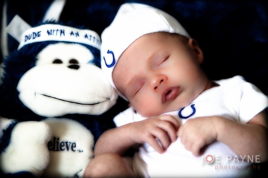 A sleeping baby wearing a white outfit and cap lies next to a stuffed monkey toy. The monkey has a headband with text and a shirt that says believe.