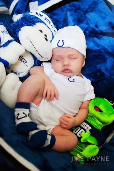 A sleeping baby wearing a white outfit with a horseshoe logo lies on a blue blanket, surrounded by a blue and white stuffed monkey and a green stuffed frog. The baby also wears matching socks and a hat with the text DUDE WITH A DO.
