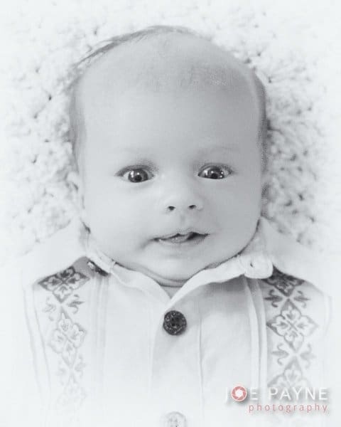 Black and white photo of a baby lying on a soft surface. The baby has wide eyes and a slight smile, wearing a patterned shirt. The background is textured.