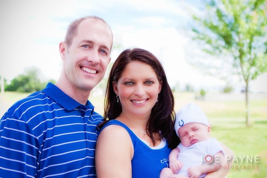 A smiling couple stands outdoors holding a sleeping baby. The man and woman are wearing blue clothes, and the baby has a white hat with a small blue horseshoe design. A tree and a bright sky are visible in the background.