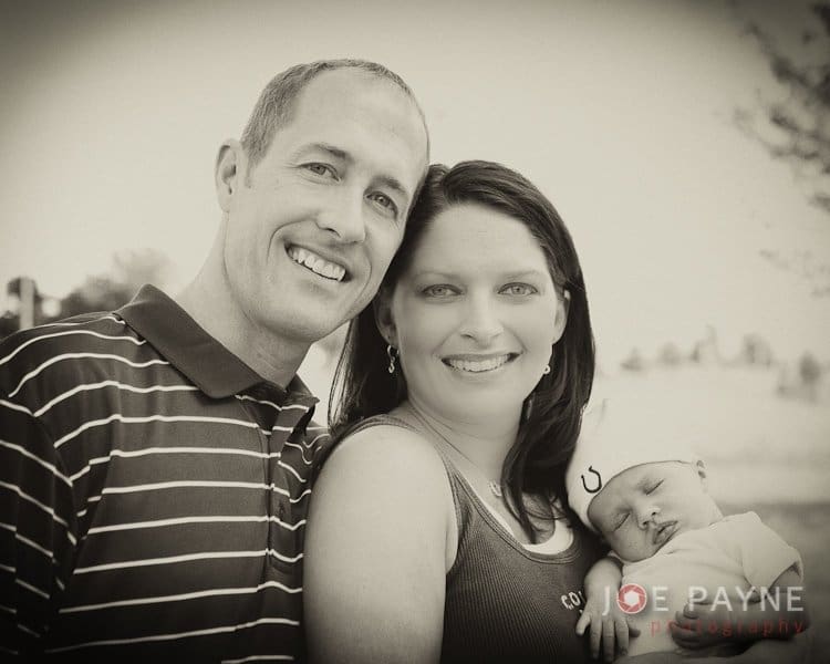 A smiling couple poses outdoors, with the woman holding a sleeping baby wearing a Colts beanie. The photo is in black and white, and they appear happy and relaxed.