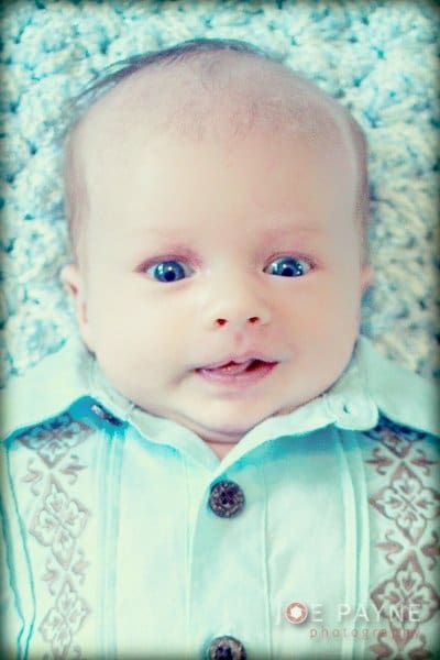 A close-up of a baby lying on a soft surface, wearing a light blue shirt with intricate patterns. The baby has wide eyes and a slight smile.