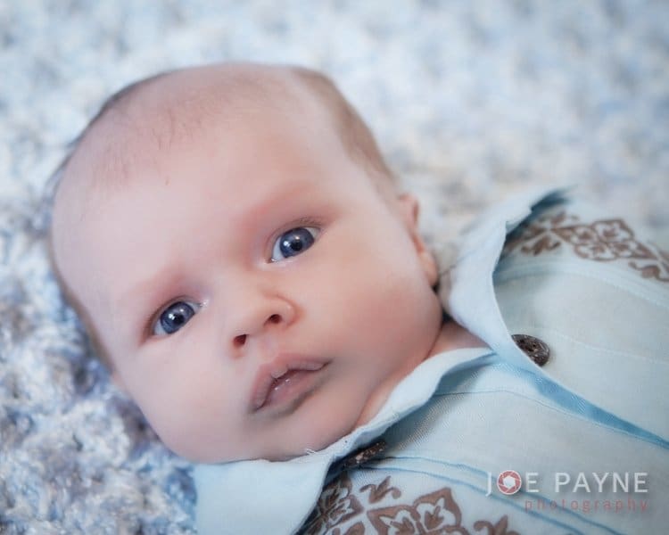 A baby with wide eyes wearing a light blue outfit with brown patterns lies on a soft, textured blanket. The image has a gentle, serene atmosphere.