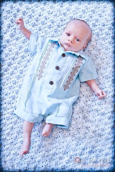 A baby in a light blue romper with decorative patterns on the front lies on a textured blue and white blanket. The babys eyes are open and one arm is slightly raised. The image has a soft, blue-tinted border.