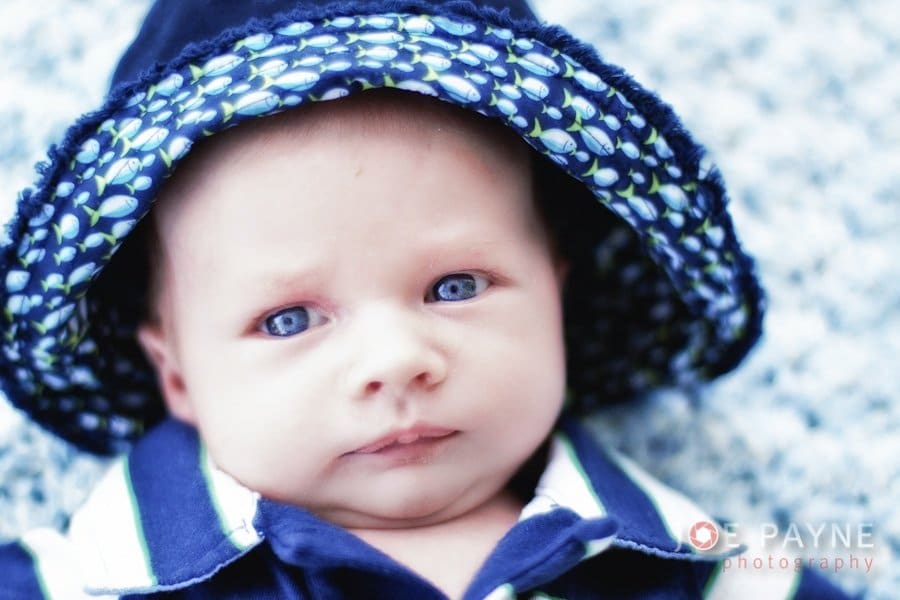 A baby with bright blue eyes, wearing a dark blue hat with a leaf pattern, gazes intently at the camera. The baby is dressed in a blue and white outfit, lying on a soft, textured surface.