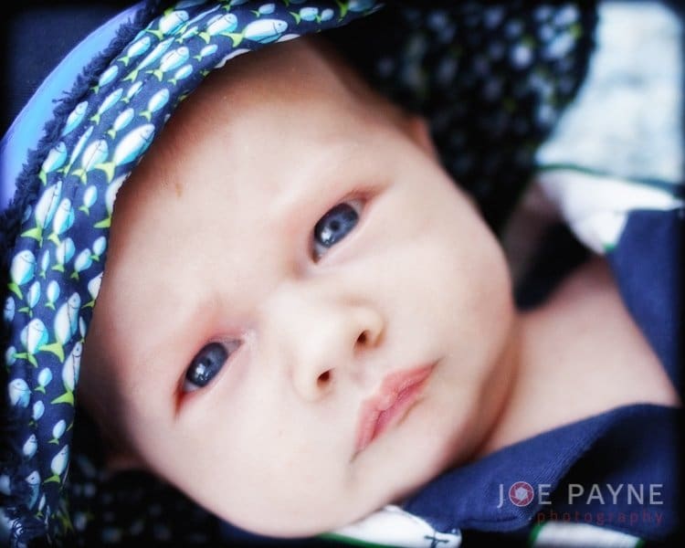 A close-up of a baby with blue eyes, wearing a patterned hat. The baby looks intently at the camera. The name Joe Payne is visible in the bottom right corner.
