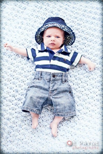 A baby wearing a striped shirt, denim shorts, and a blue sun hat lies on a soft, textured blanket. The baby looks up with wide eyes, arms stretched out to the sides.