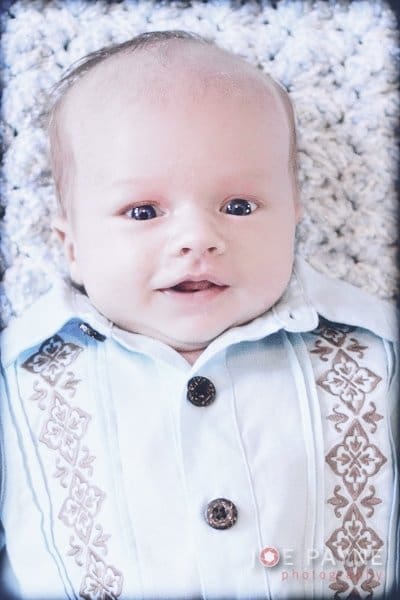 A baby in a light blue shirt with embroidered patterns on the collar and chest is lying on a textured light-colored blanket, looking up with a slight smile.