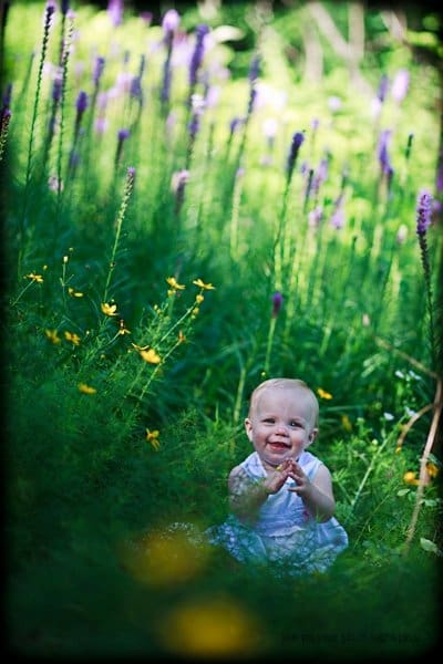 A baby in a white dress sits smiling in a lush green field, surrounded by tall purple and yellow wildflowers, under bright sunlight.