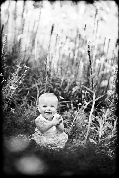 Black and white photo of a smiling baby sitting in tall grass and wildflowers. The baby wears a light-colored sleeveless outfit and looks directly at the camera, surrounded by a natural, blurred background.