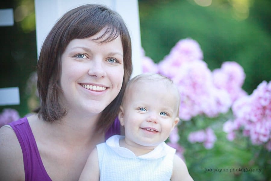 A smiling woman with short brown hair holds a happy baby wearing a white outfit. They are outdoors, surrounded by blooming pink flowers and greenery.