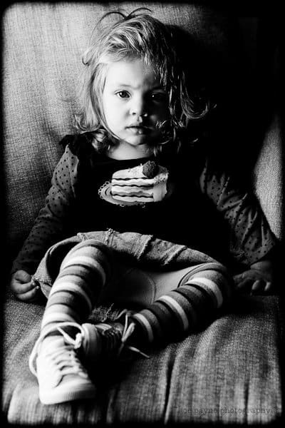 Black and white photo of a young girl sitting on a chair, looking at the camera as if catching up with an old friend. She has messy hair, wears a cake design shirt, striped leggings, and sneakers. Light and shadow highlight her face.