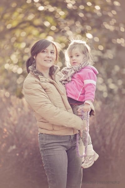 A woman in a tan jacket holds a young girl in a pink jacket outdoors, with soft sunlight and blurred trees in the background. Both look toward the camera, and the mood is warm and gentle.
