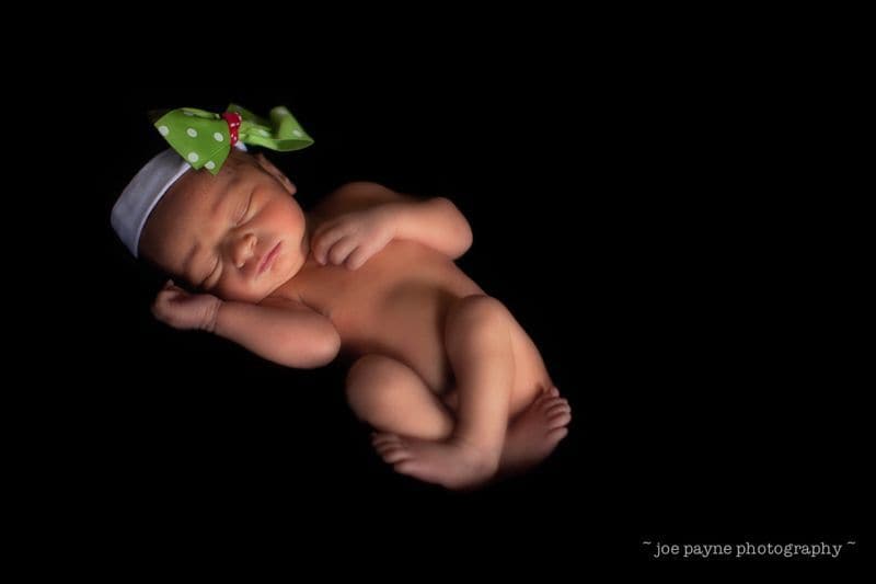 A newborn baby sleeps curled up on a black background, wearing a white headband with a large green bow decorated with white polka dots. With soft, gentle lighting, the scene perfectly captures the magic moment when a star is born.