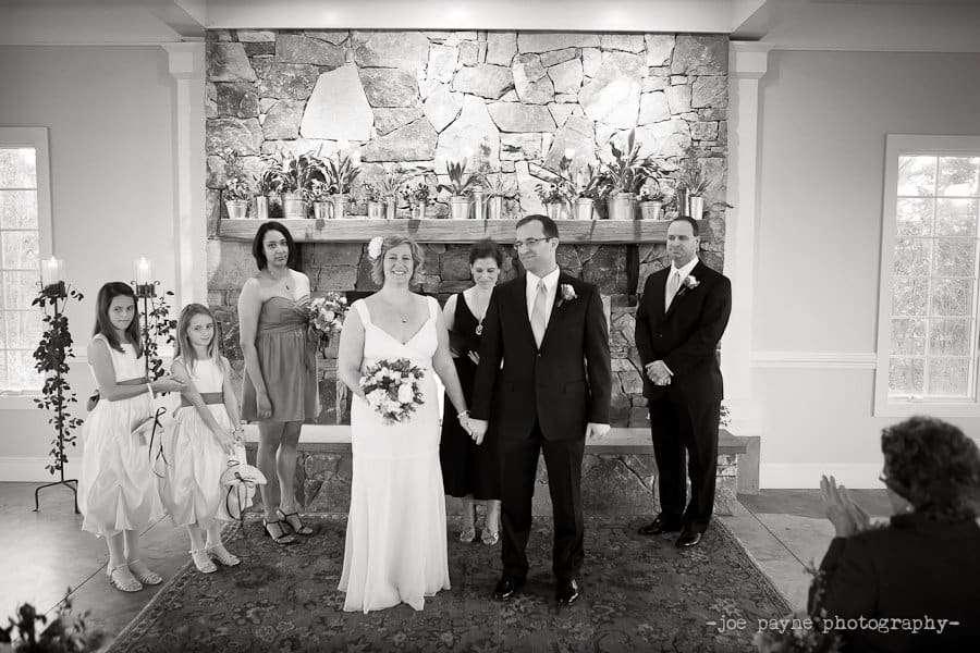 A bride and groom are standing hand in hand in front of a stone fireplace during a wedding ceremony. Bridesmaids and a groomsman stand beside them. A seated guest claps in the foreground. The scene is elegant and serene.